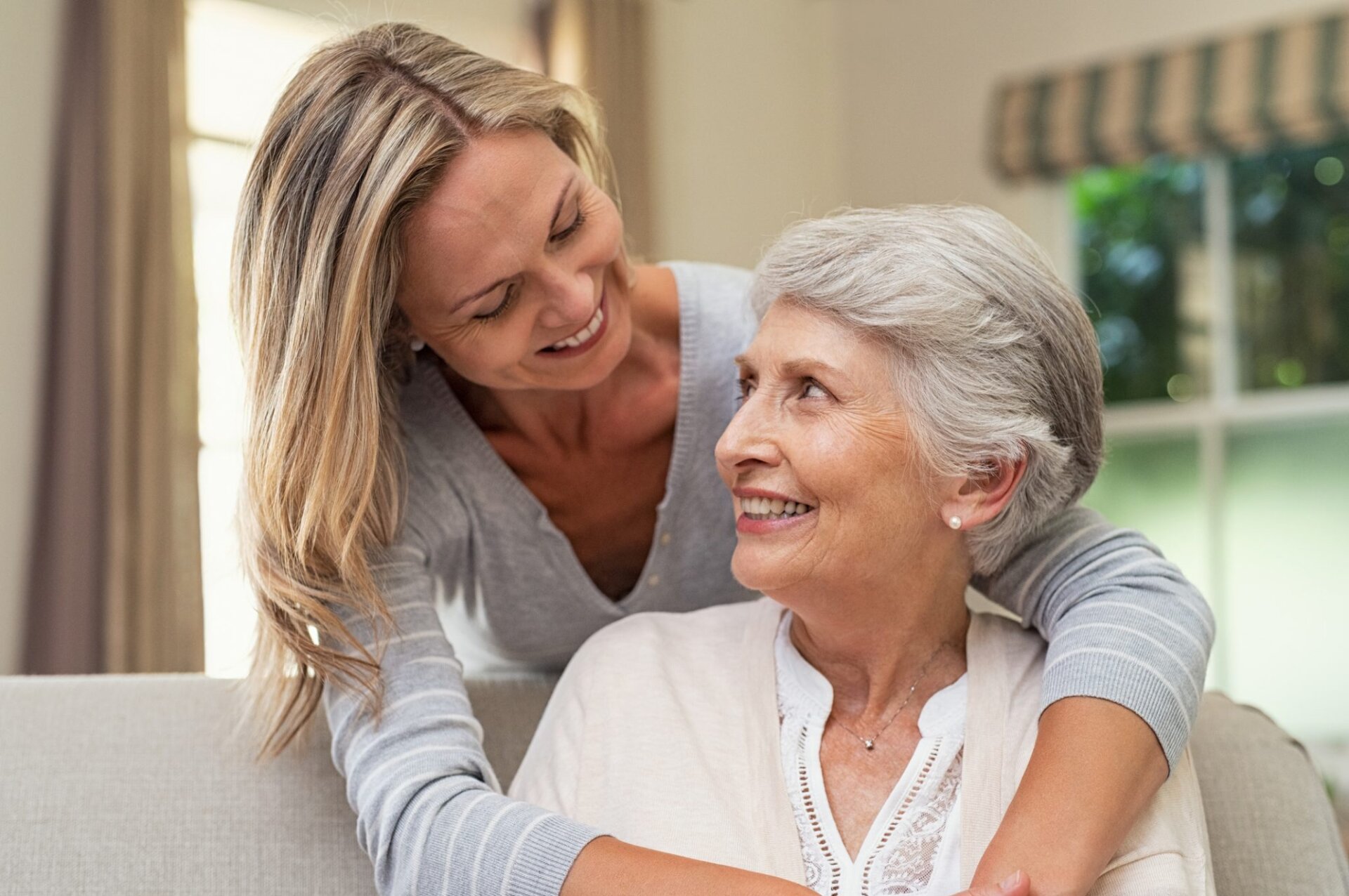 Senior woman sitting on couch while woman hugs her from behind