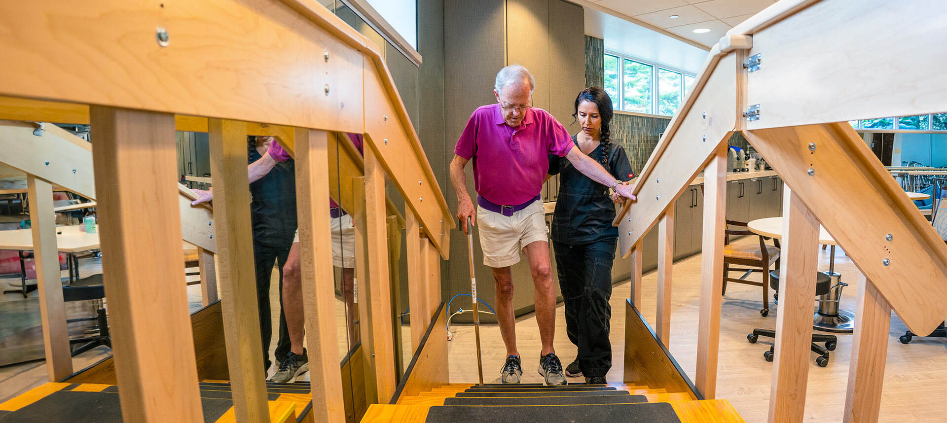 Senior man receiving assistance from a caregiver while walking up stairs in a common area.