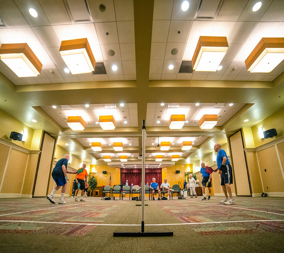 Group of seniors playing indoor badminton in a well-lit communal area with high ceilings.