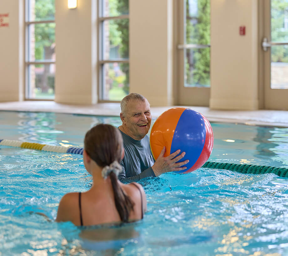 Two individuals enjoy a swimming pool in a senior living community, playing with a beach ball.
