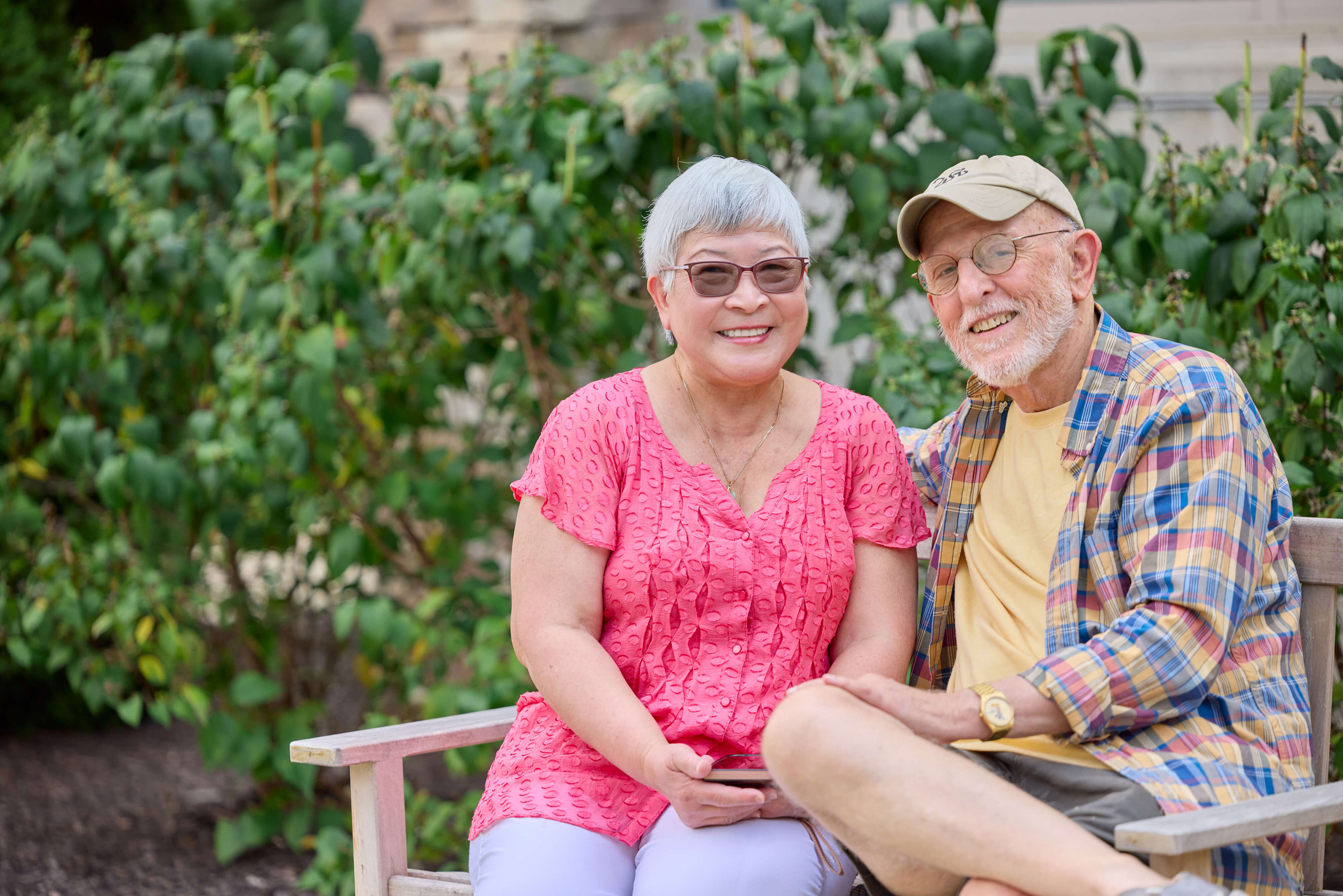 Two senior residents sitting on a bench smiling in a green courtyard in front of a living unit.