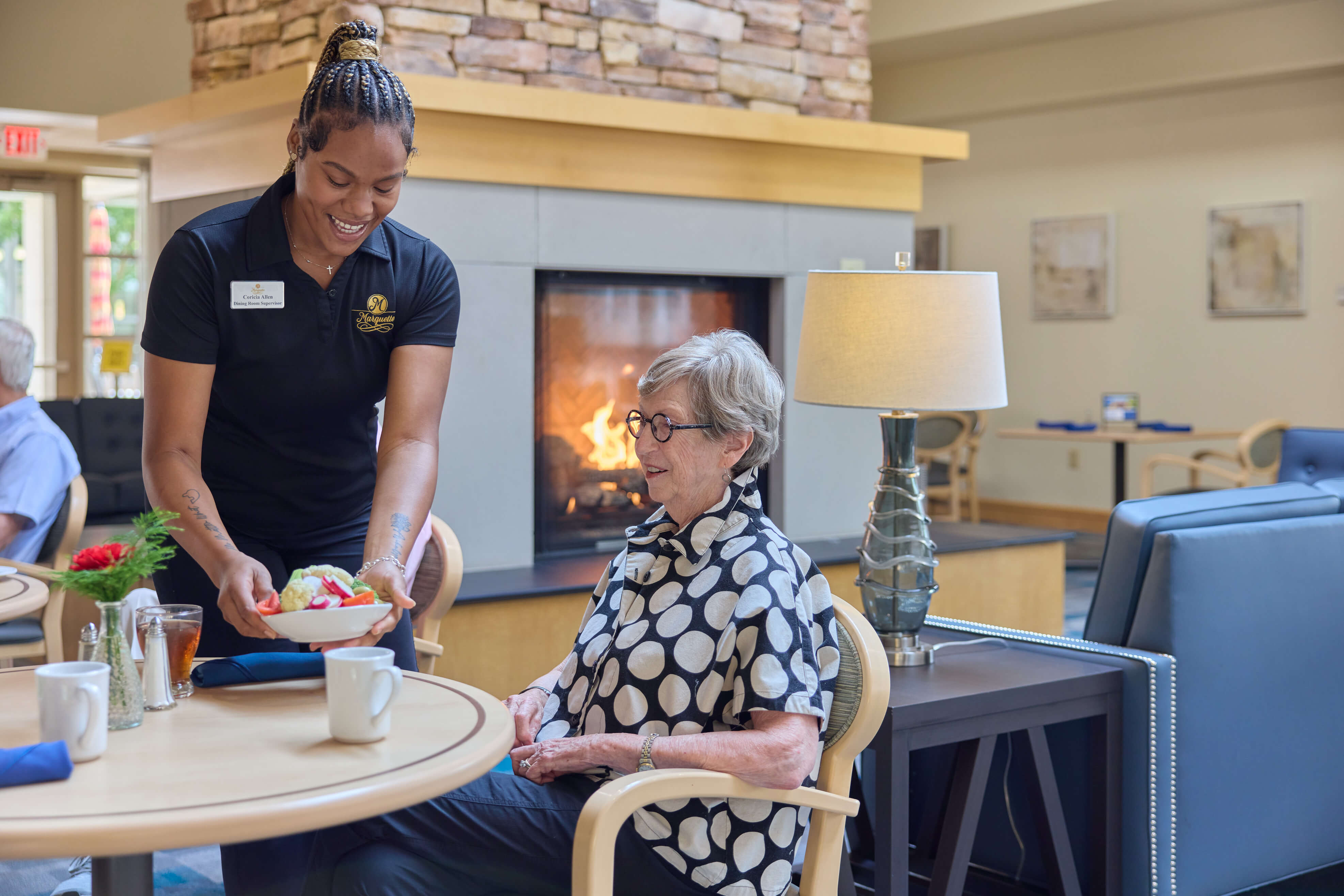 Staff member serves a meal to a seated resident at a senior living community dining area.