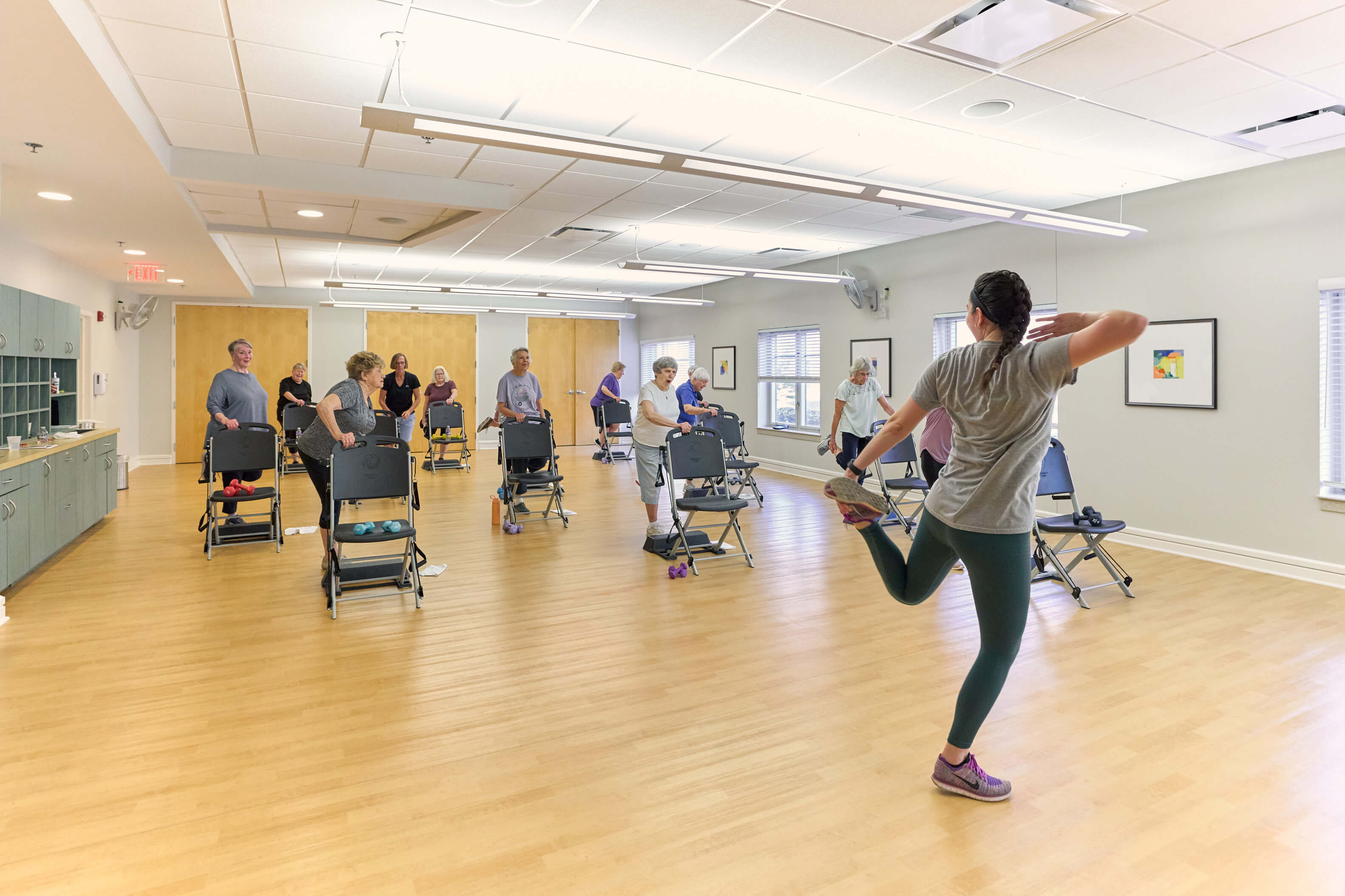 Group exercise class with participants in a well-lit room using chairs for support.