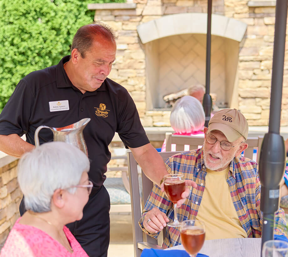 Staff member serves drink to a seated resident in outdoor patio of a living community.