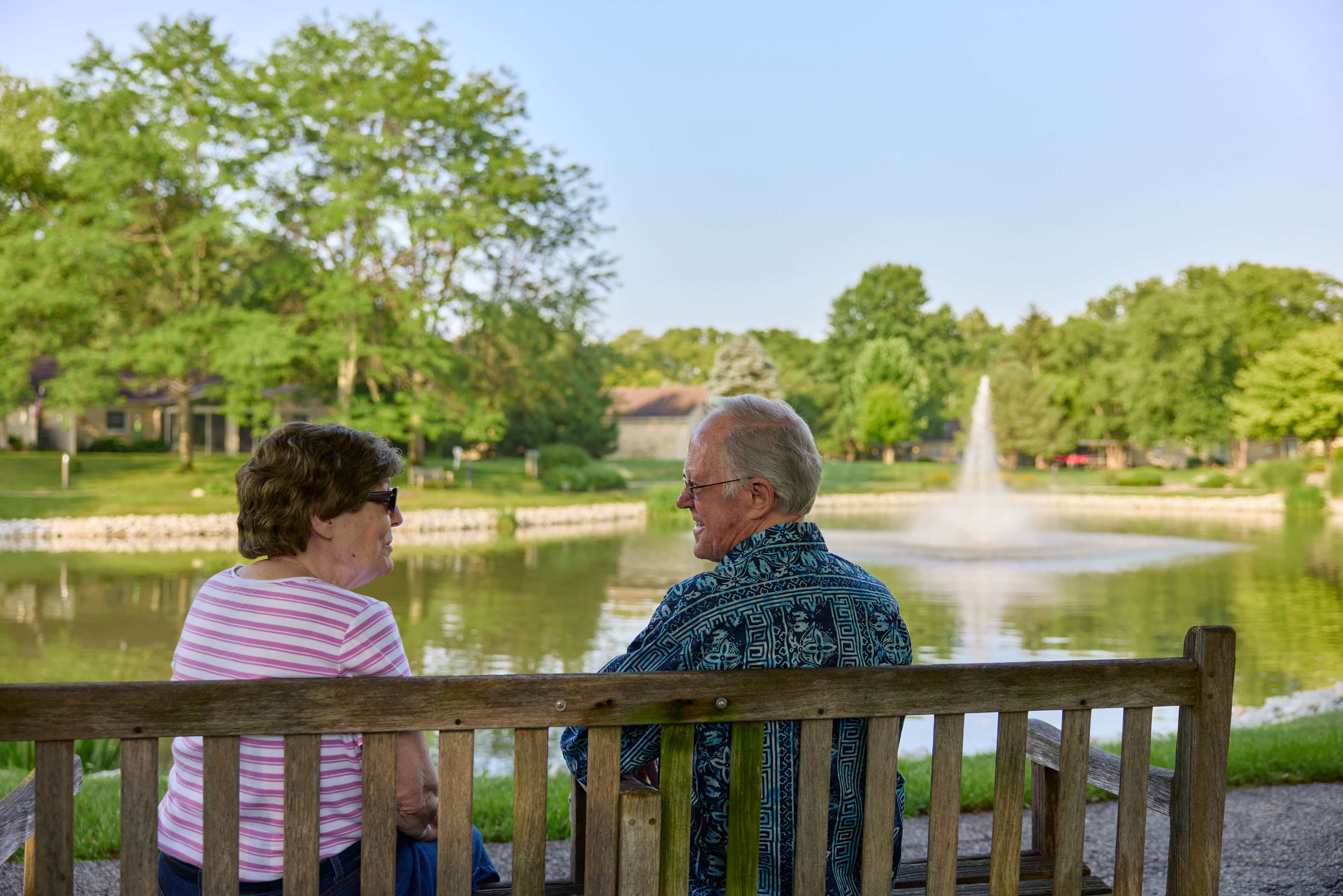 Two seniors sitting on a bench by a pond with a fountain at Marquette garden area.