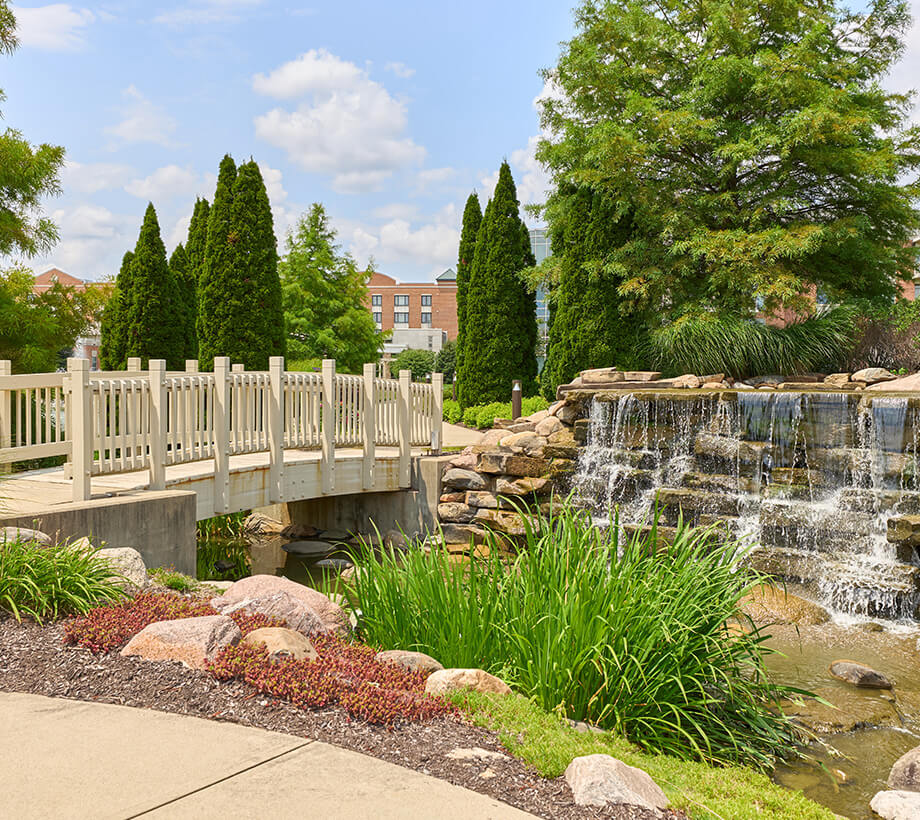 Scenic outdoor area with a white bridge, waterfall, and lush greenery in a senior living community.