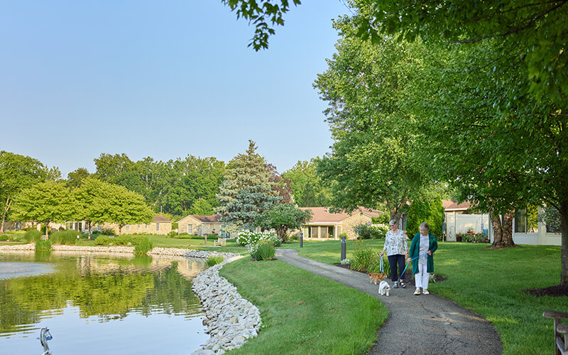 Two women walking a small dog on a pathway beside a pond with surrounding greenery.