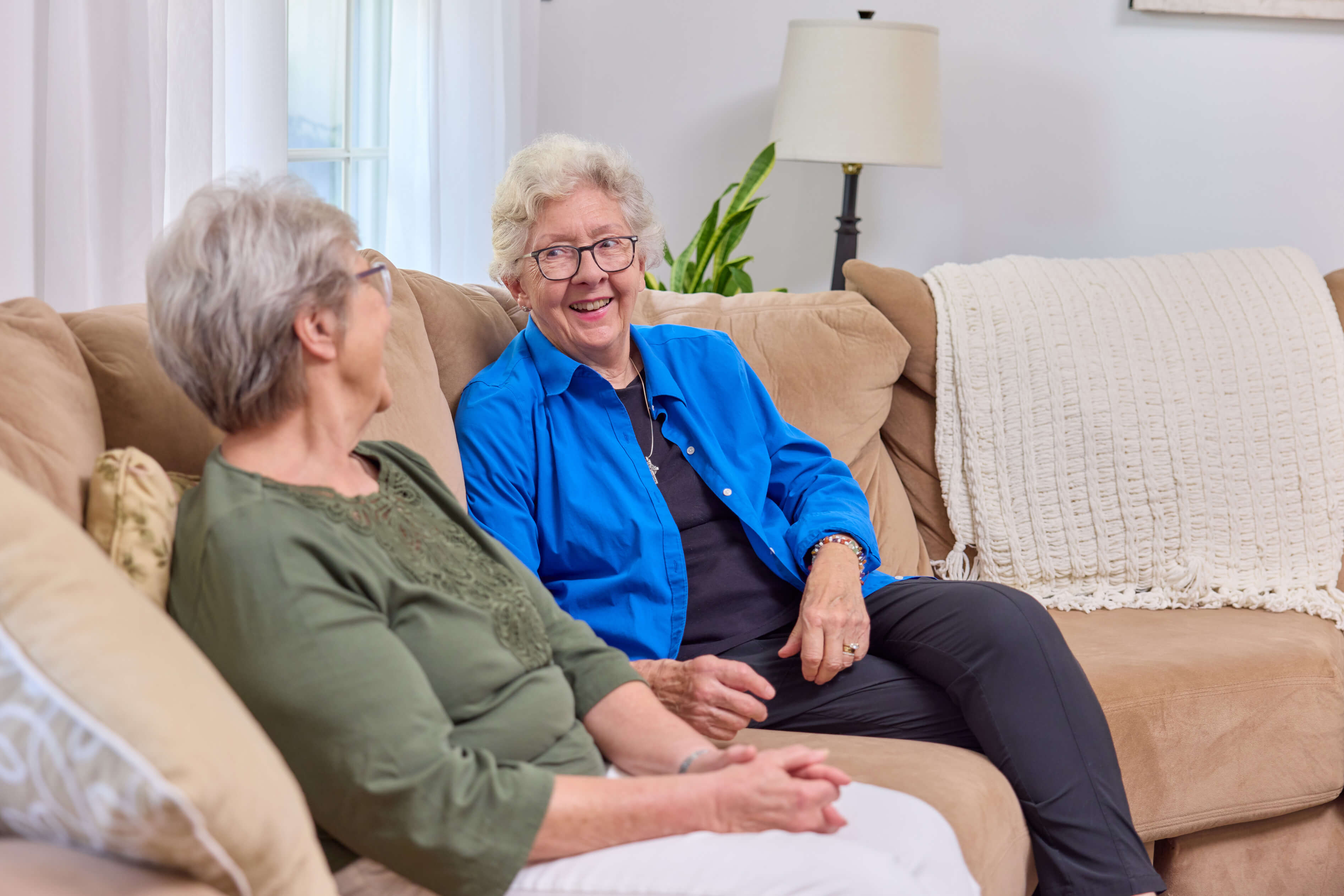 Two elderly women sitting on a beige sofa, having a conversation in a cozy living space.