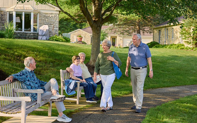 Elderly residents strolling and sitting on a bench in a green, tree-lined walkway near units.