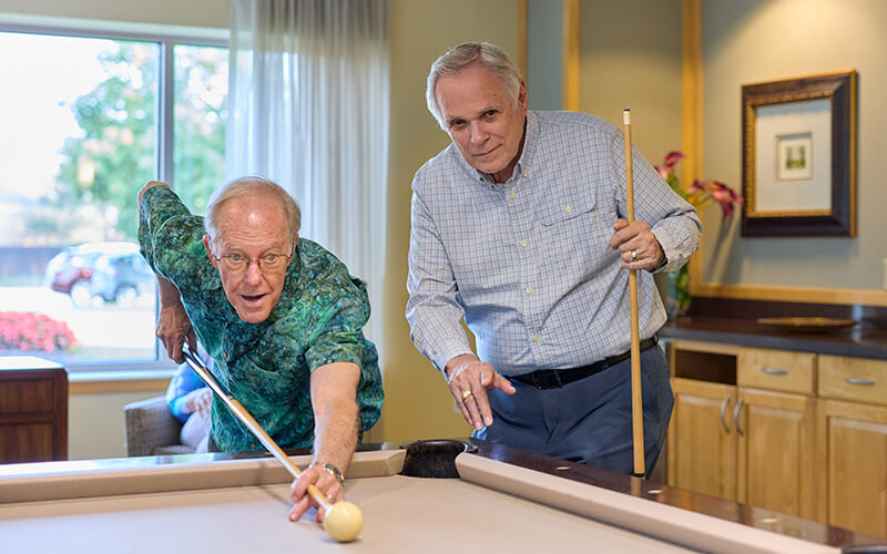 Two senior men playing billiards in a bright, cozy community recreation room.