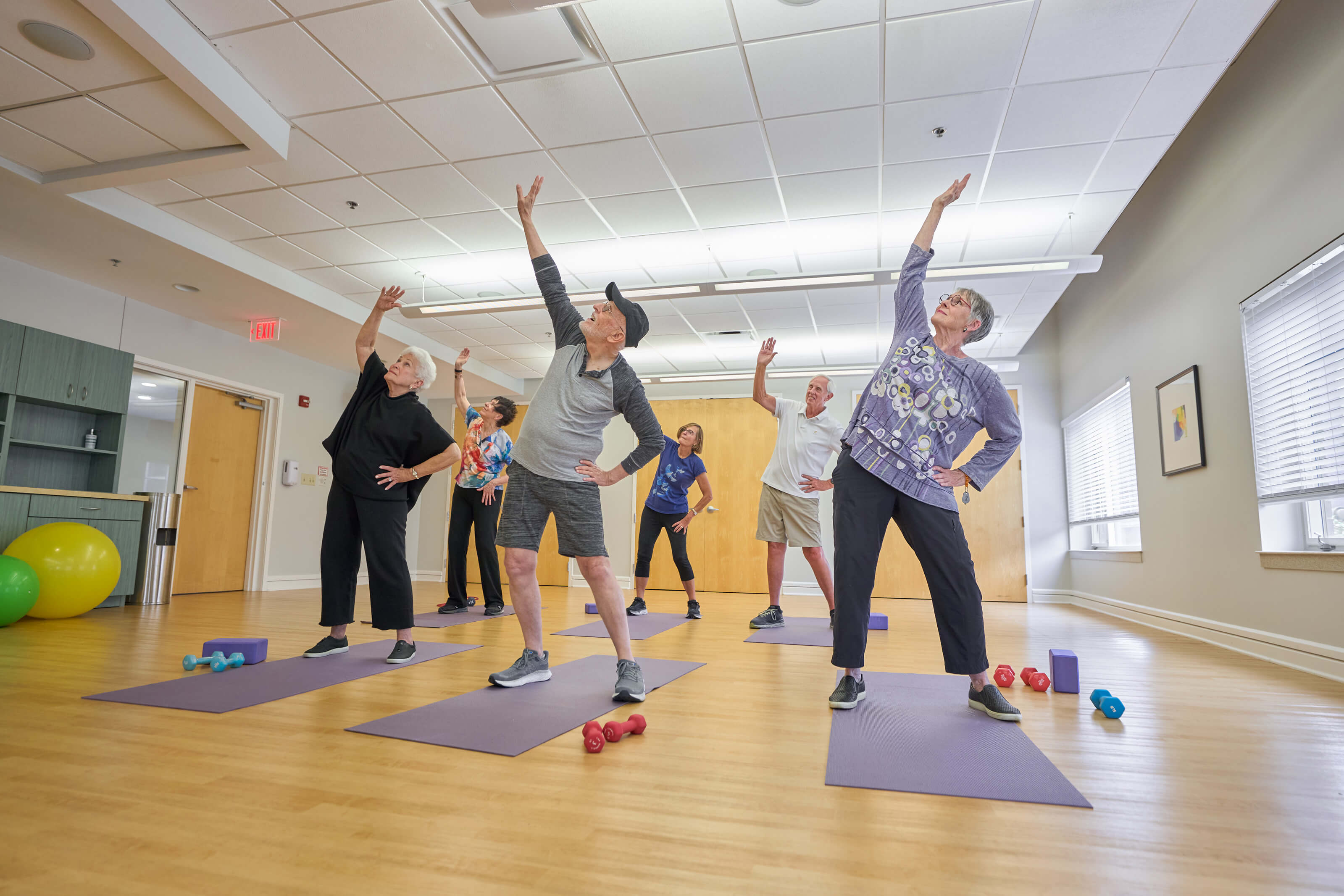 Group of seniors exercising in a well-lit room with wooden floors and exercise equipment.