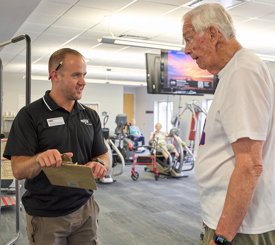 Fitness trainer with clipboard talking to senior man in gym area of a senior living community.