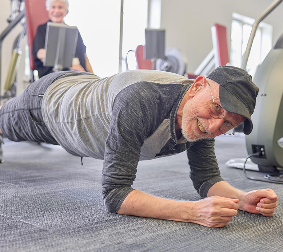 Senior man smiling while doing a plank exercise in a gym setting with another man using gym equipment in the background.