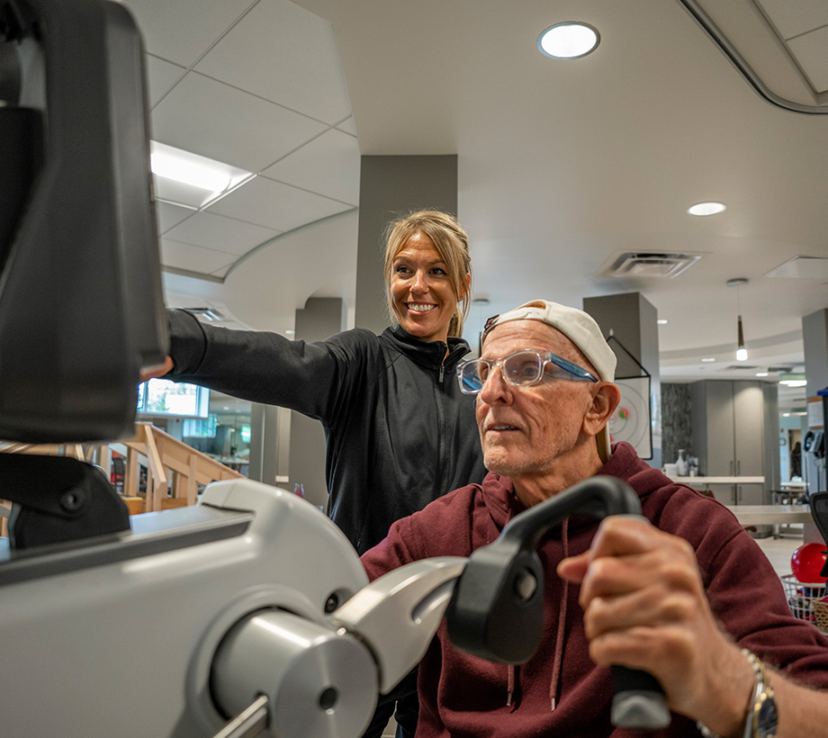 Senior man exercising on gym equipment while a trainer assists him in the community fitness center.