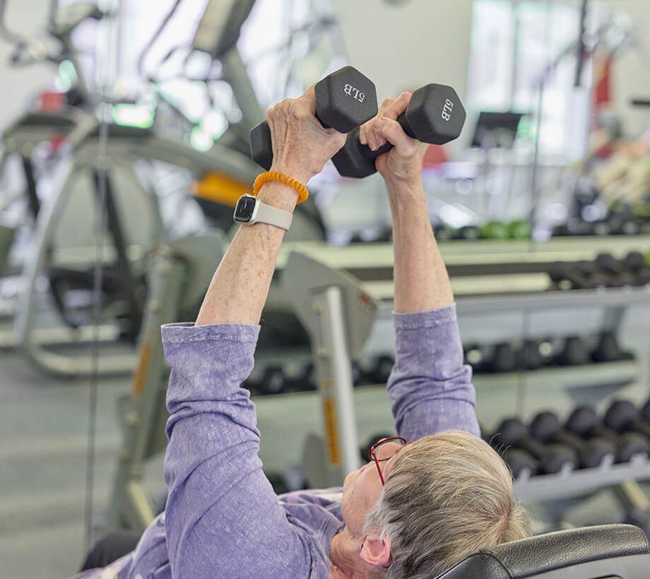 Senior woman lifting weights in a modern exercise room with various gym equipment.