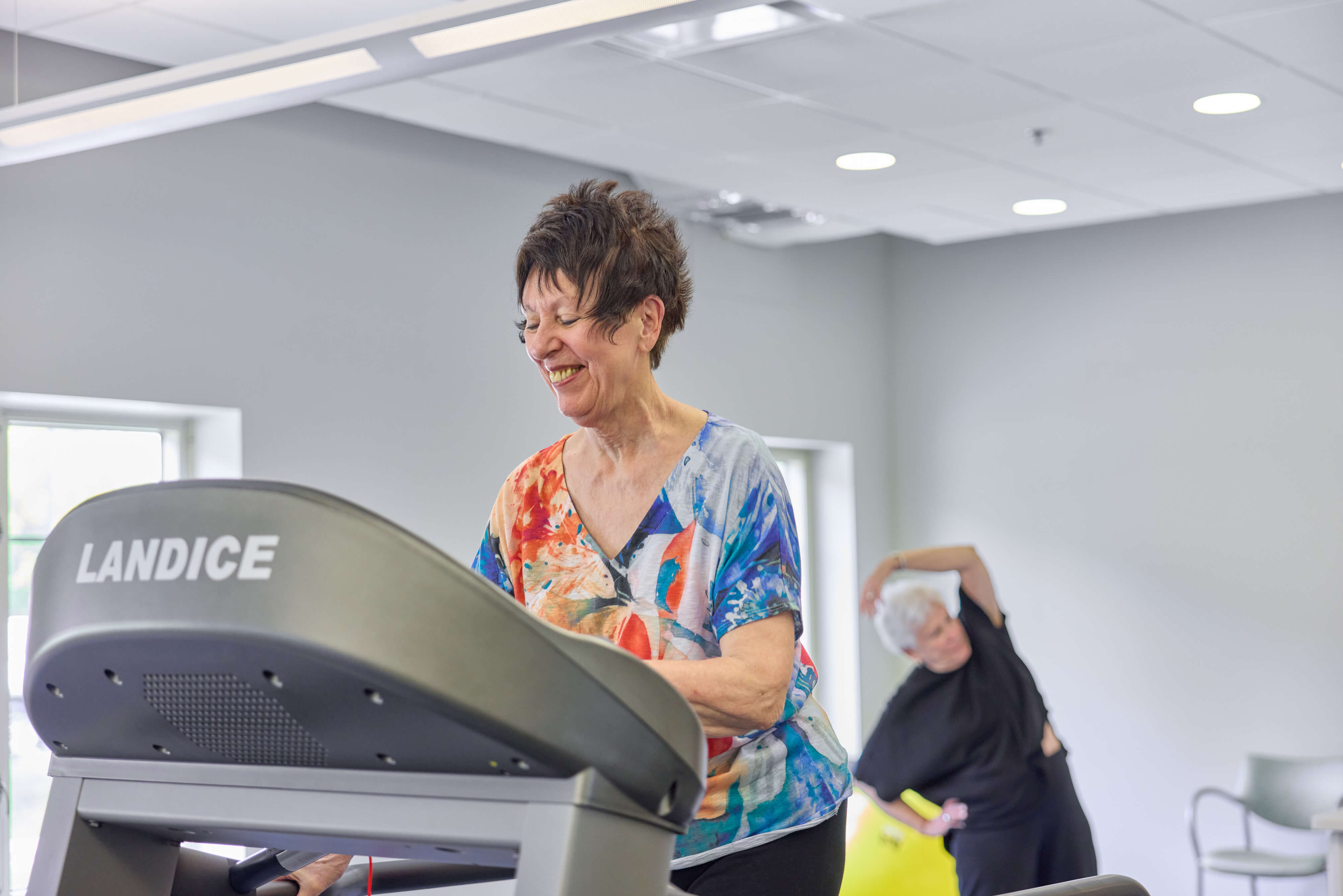 Two seniors exercising in a well-lit gym, one on a treadmill and another stretching.