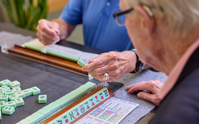 Two senior residents playing a game of Mahjong at a community center table.
