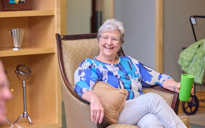 Smiling elderly woman sitting in a chair and holding a green cup in a senior living unit.
