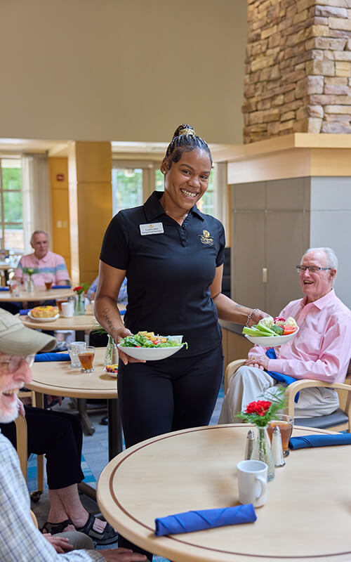 Smiling staff member serves salads to senior residents in a well-lit, communal dining area.