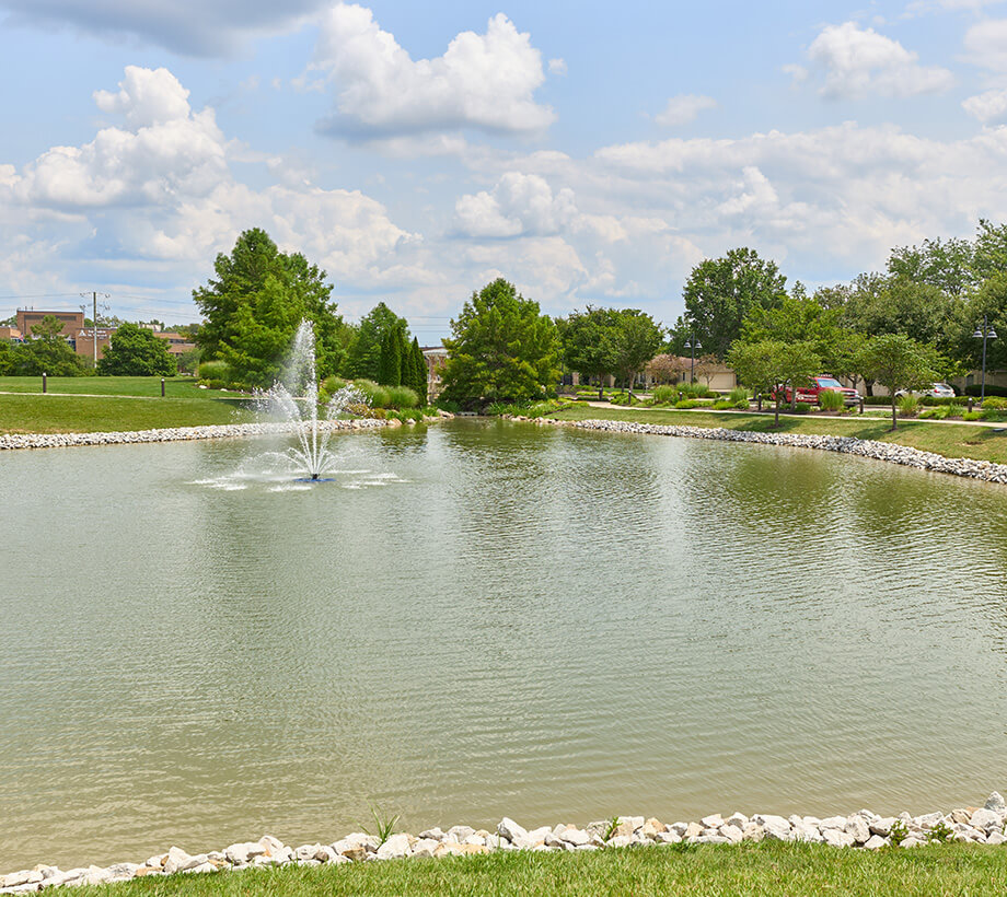 Tranquil pond in a senior living community with a central fountain and surrounding greenery.