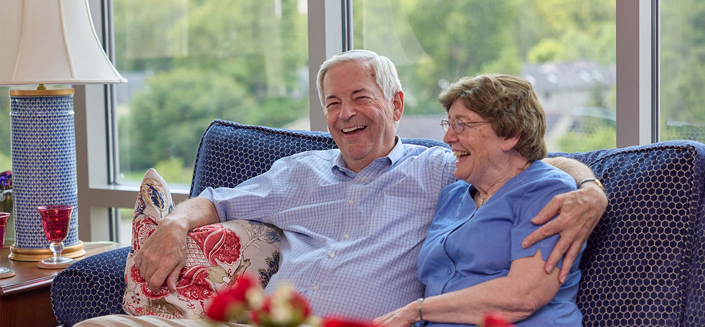 Senior couple seated on couch smile while embracing each other