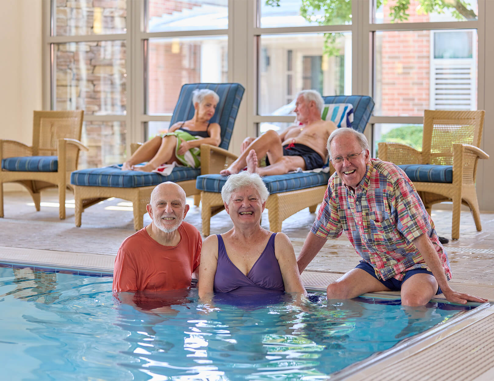 Three seniors enjoying an indoor pool while two others relax on loungers in a senior living community.