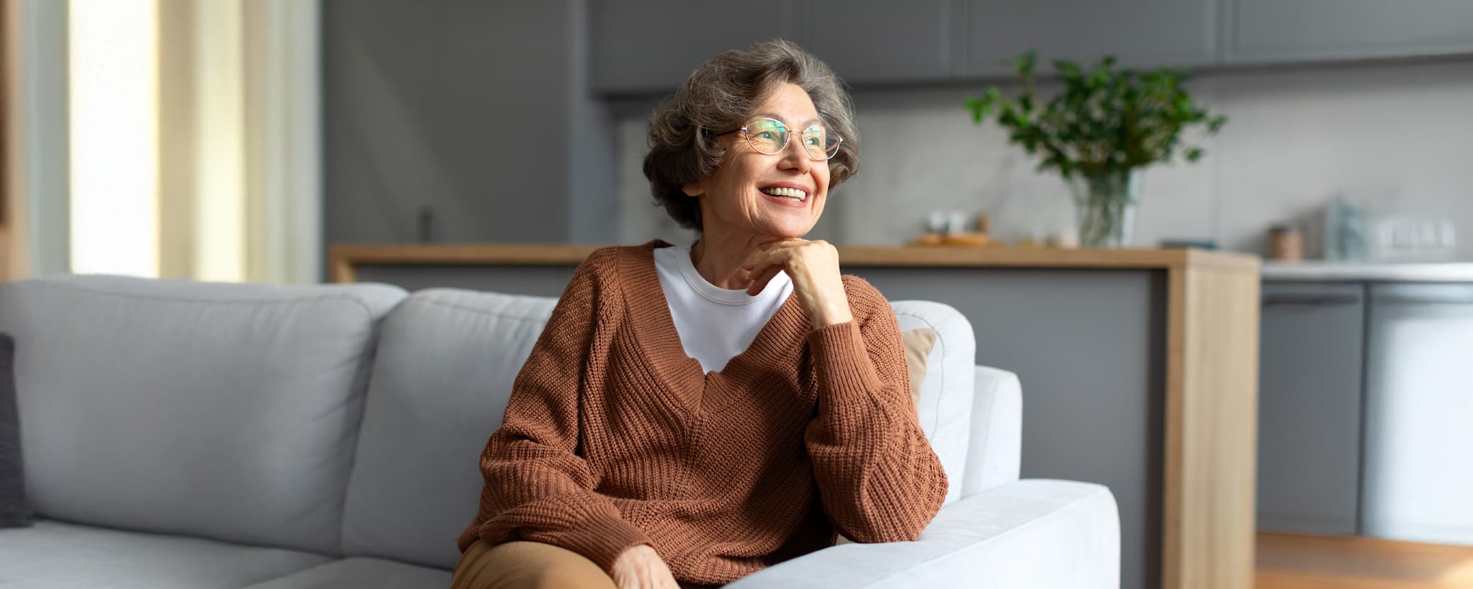 Senior woman smiles as she looks out the window from her couch