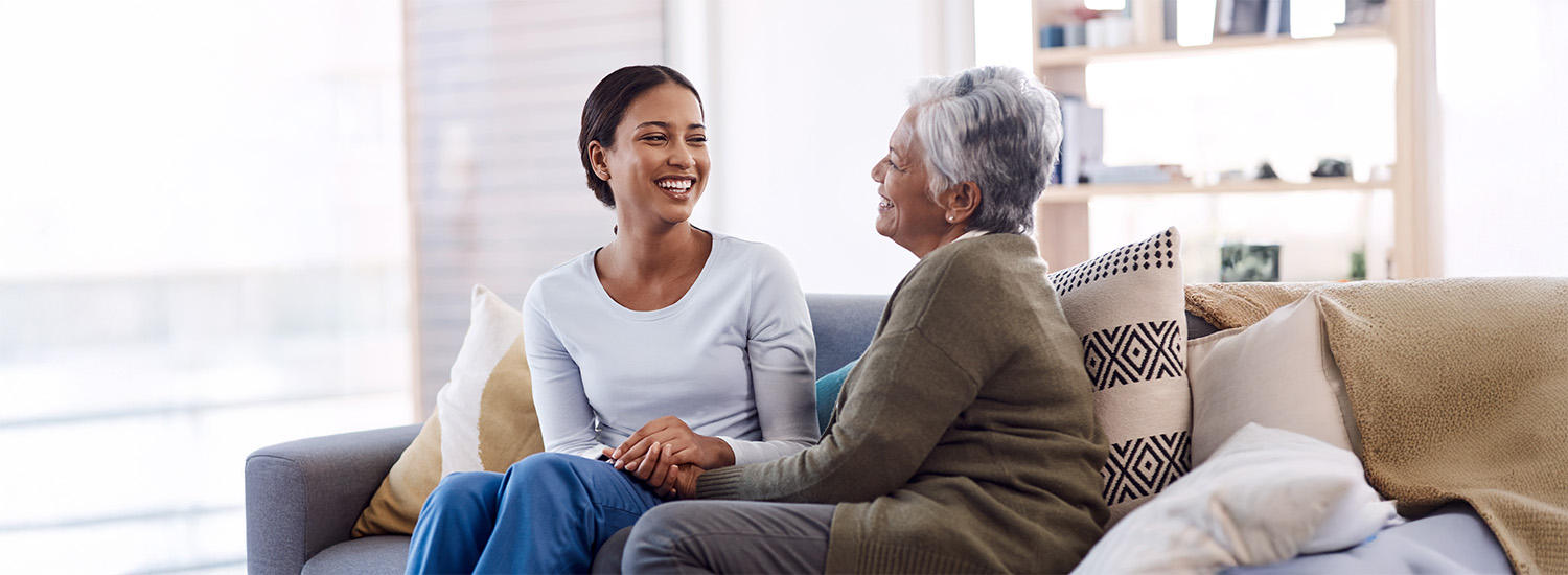Young woman sits on couch with senior woman and holds her hand while smiling at her