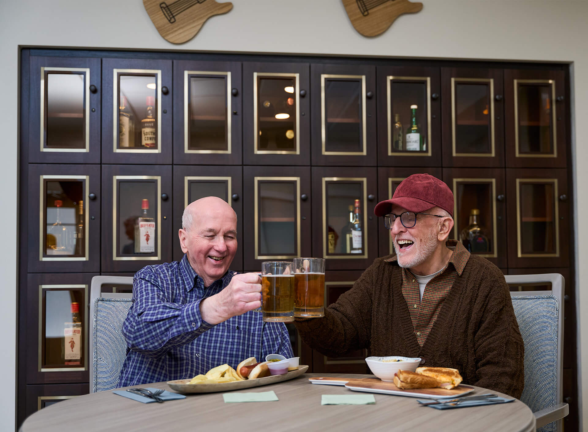 Two senior men enjoy drinks and food, smiling in a lively community lounge.