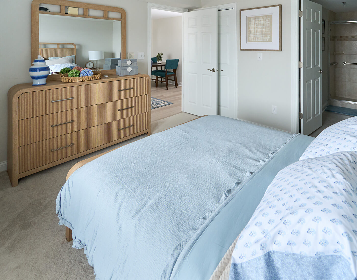 Cozy bedroom with blue bedding, wooden dresser, and an open door to another room.