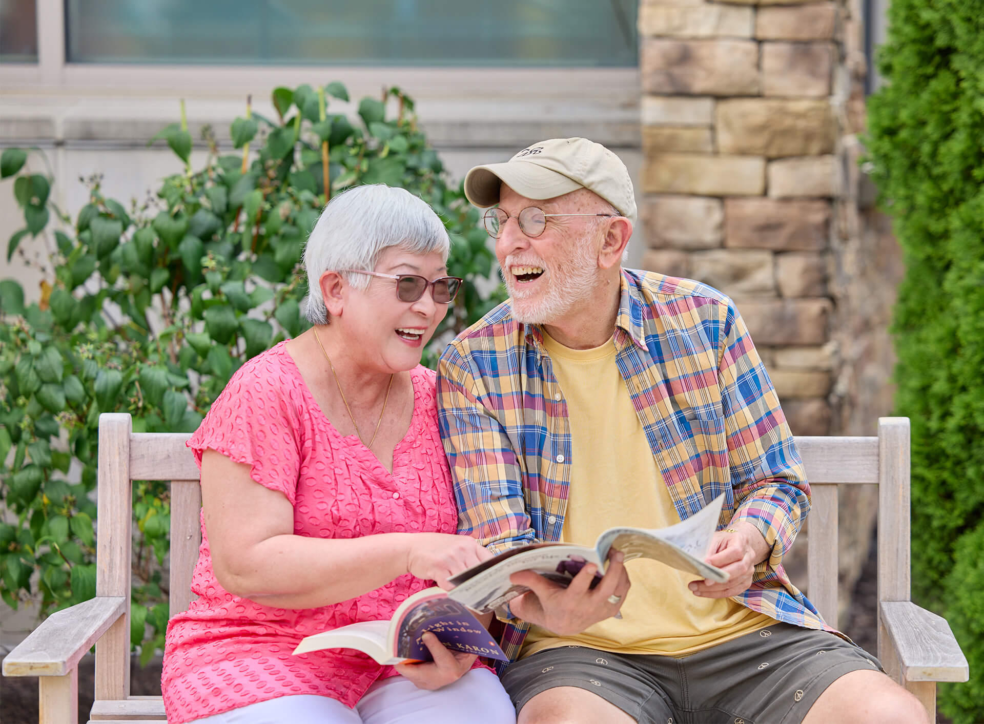Elderly couple reading and laughing on outdoor bench with plants in background.